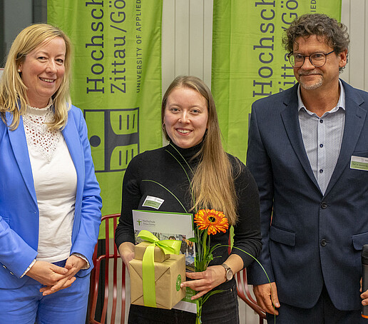 The Vice President, the Al-Makhawi prizewinner and Prof. Zips stand smiling next to each other in front of two green HSZG flags and smile into the camera.