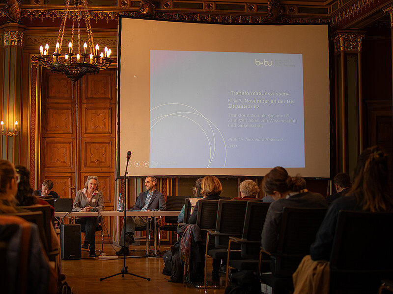 Speakers sit at tables in front of packed rows of chairs in the citizens' hall of Zittau town hall. A beamer projection on a screen shines in the background.