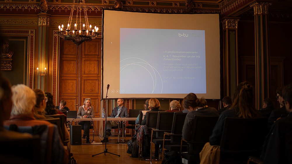Speakers sit at tables in front of packed rows of chairs in the citizens' hall of Zittau town hall. A beamer projection on a screen shines in the background.