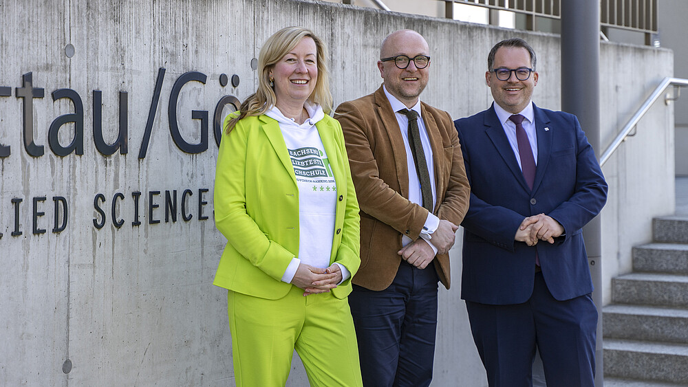 Robert Denk, Sophia Keil and Alexander Kratzsch stand in front of the HSZG logo and smile at the camera.