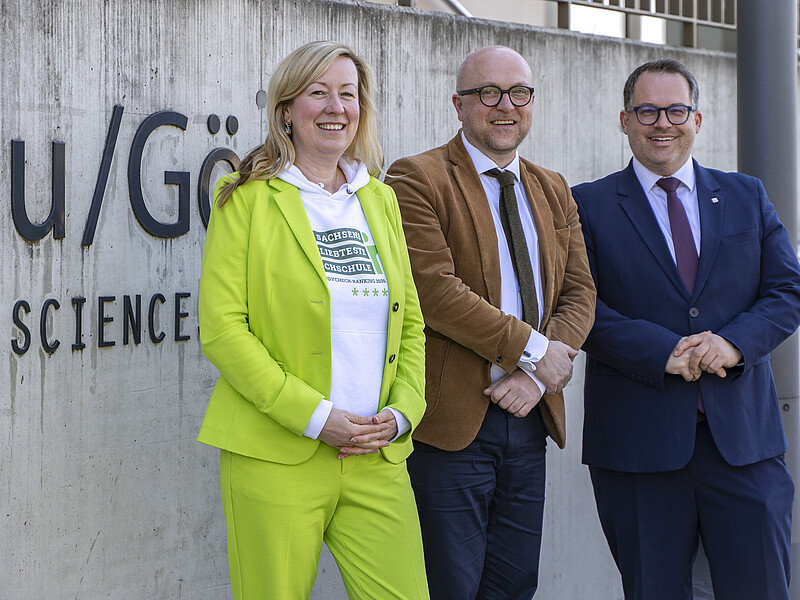 Robert Denk, Sophia Keil and Alexander Kratzsch stand in front of the HSZG logo and smile at the camera.