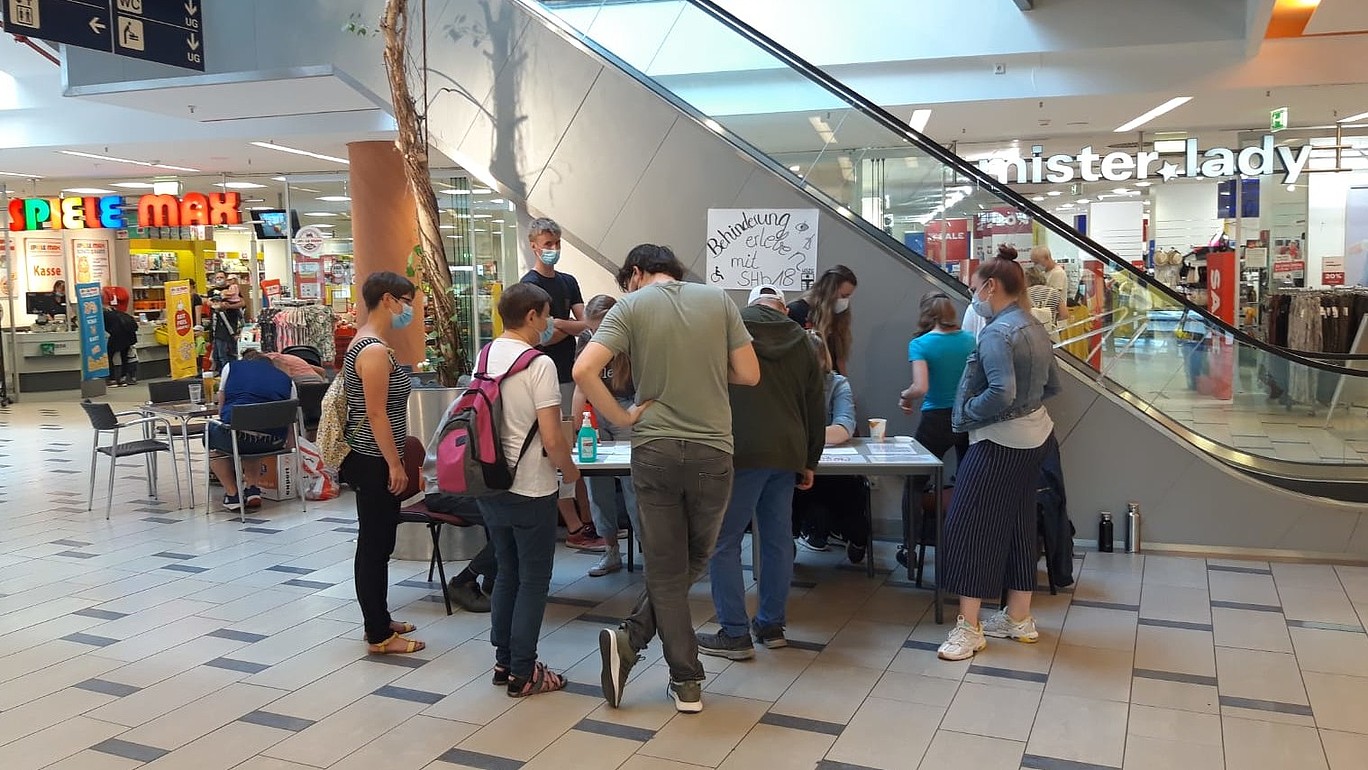 Eine Gruppe interessierter Menschen stehen im City Center vor dem Tisch, um Behinderung zu erleben.