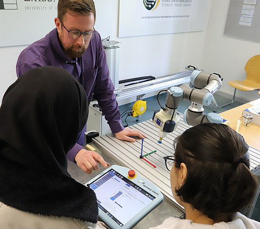 Christian Vogel points to a screen at which two schoolgirls are looking. The cobot is standing on a table next to it.