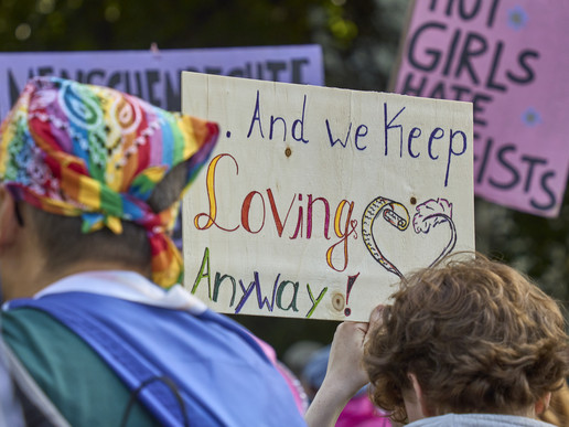 Ein CSD-Teilnehmer mit Regenbogenfarbigen Kopftuch und eine Begleitung laufen beim Christopher Street Day mit. Man sieht ihre Rückenansicht. Dazwischen ist ein Schild mit der Aufschrift: And we keep loving anyway, zu lesen.