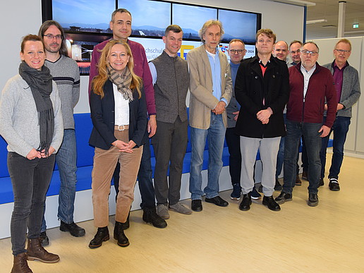 A group of HSCG employees and cooperation partners stand in front of the monitor wall in the laboratory hall.