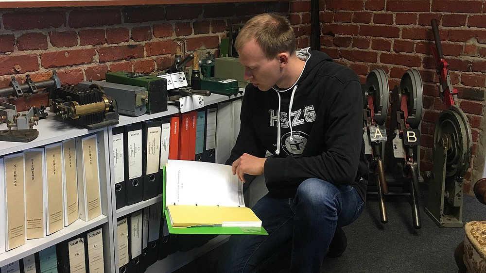 A man squats in a room in front of a shelf with many folders and holds a folder in his hand in which he reads something
