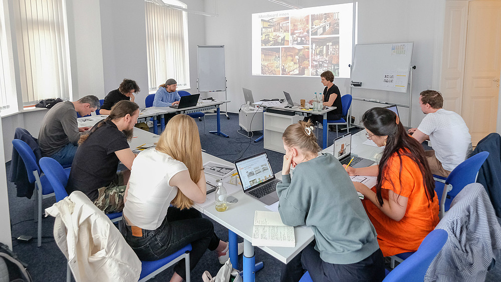 A group of course participants are sitting in a seminar room and taking notes. The lecturer sits at the table and explains the course content using the beamer projection.