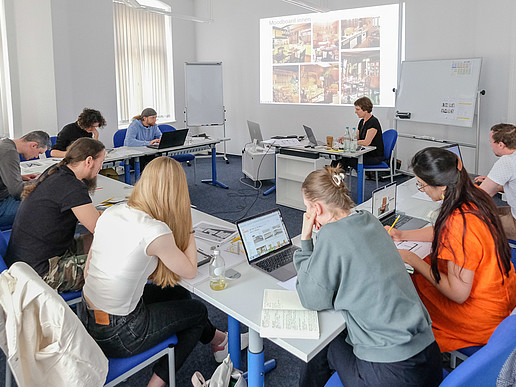 A group of course participants are sitting in a seminar room and taking notes. The lecturer sits at the table and explains the course content using the beamer projection.