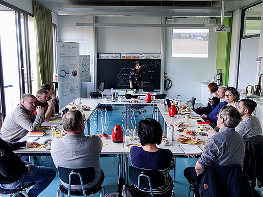 Die Teilnehmer*innen sitzen zusammen an einem Tisch vor einer Tafel und unterhalten sich.an einem 