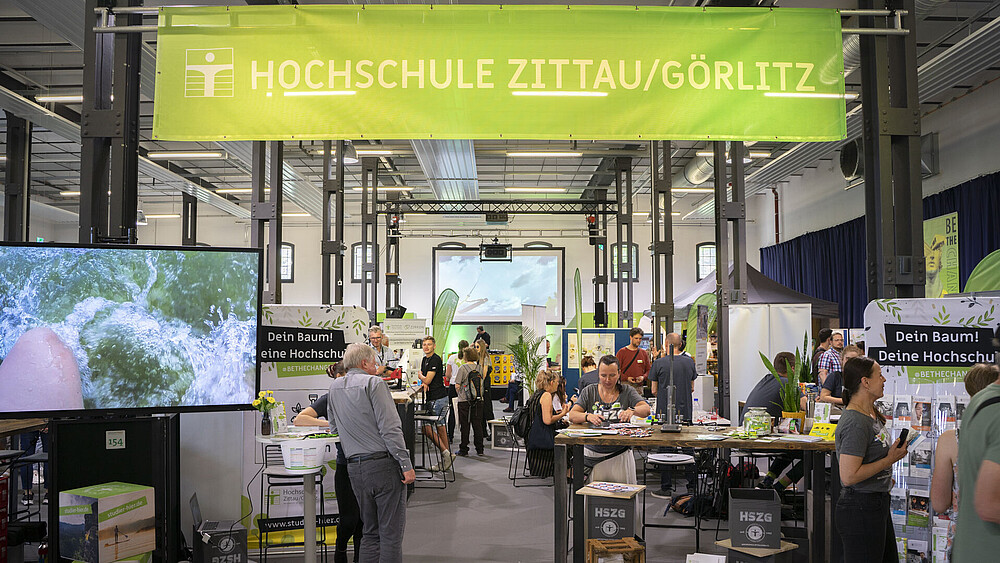 Verschiedene Versuchsstände mit Experimenten sind in einer Halle aufgebaut. Darüber ist ein Banner mit der Aufschrift Hochschule Zittau/Görlitz angebracht.