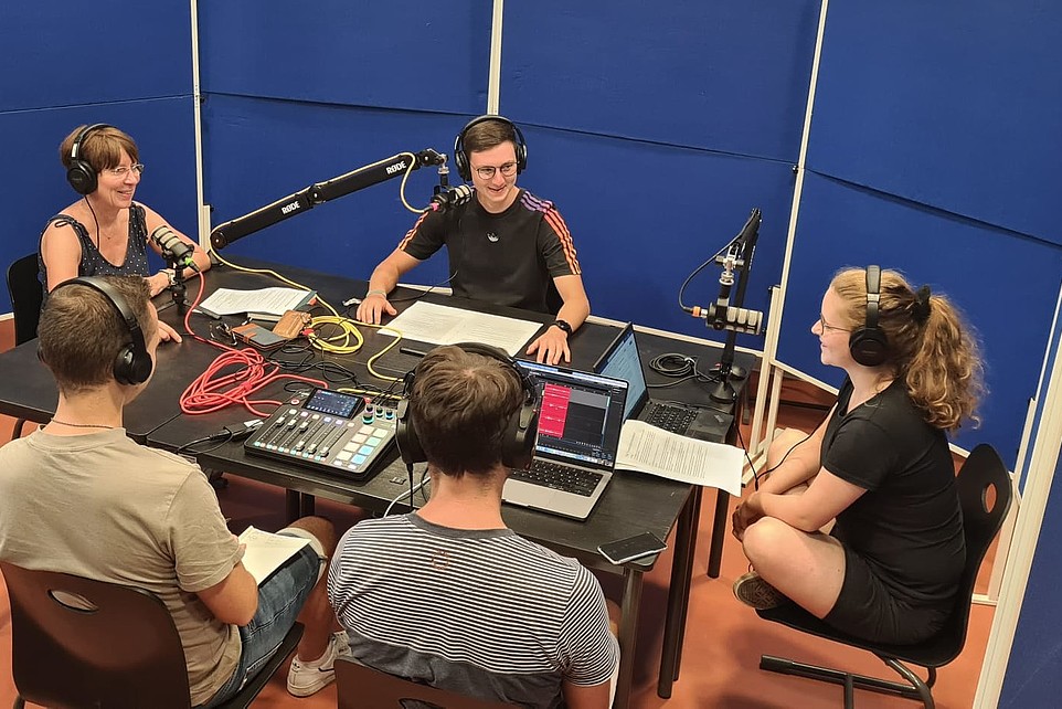 Podcastteilnehmende sitzen am Tisch umgeben von Schallwänden. Das Aufnahmeequipment steht auf dem Tisch.