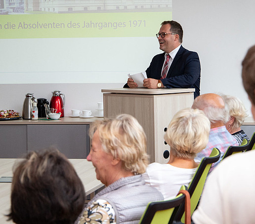 Der Rektor steht am Rednerpult im Senatssaal und hält vor älteren ehemaligen Studierenden eine Rede. Er lächelt sie freundlich an.