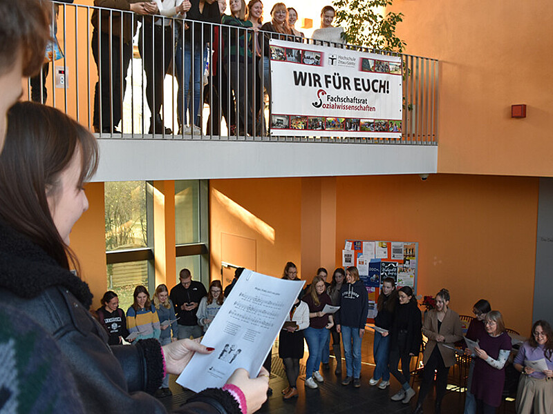 Several people are standing in the stairwell of a campus building, holding sheet music and singing.