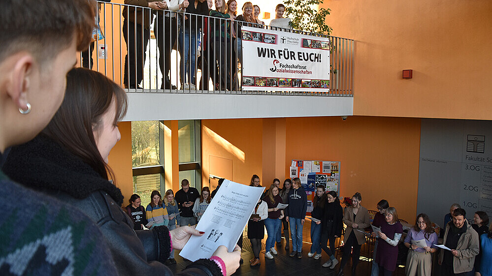 Several people are standing in the stairwell of a campus building, holding sheet music and singing.