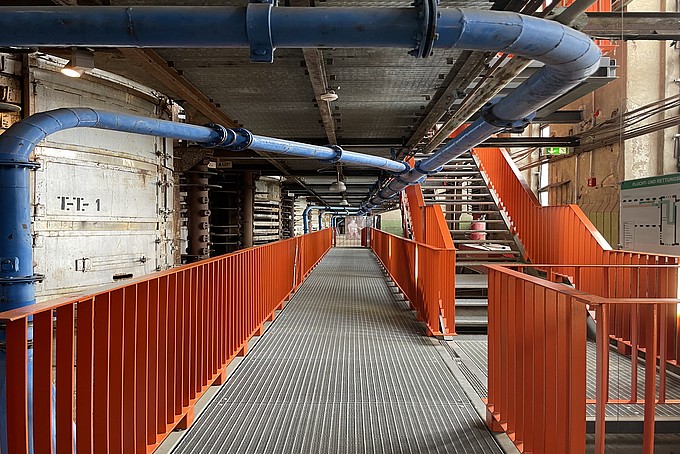Ein Gittergang in einem Gebäude der Energiefabrik mit orangenem Geländer, an der Decke verlaufen dicke blaue Rohre.