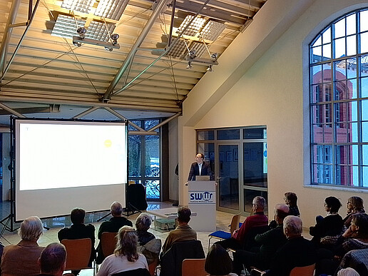 Senior citizens sit on rows of seats in front of a screen in the E-Werk. Prof. Kunick stands behind a lectern.