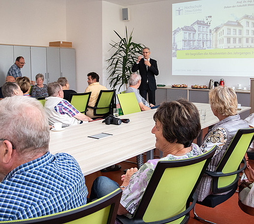 Ältere ehemalige Studierende sitzen an einer Tafel im Senatssaal. Prof. Bellair steht im Hintergrund und unterhalten sich mit ihnen.