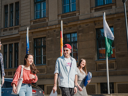 A group of international students walk across the campus laughing. European flags are waving in the background. They are interested in International Business Communication.