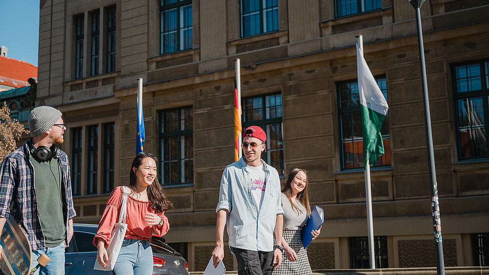 Eine Gruppe internationaler Studierender läuft lachend über den Campus. Im Hintergrund wehen europäische Flaggen. Sie interessieren sich für Internationale Wirtschaftskommunikation.