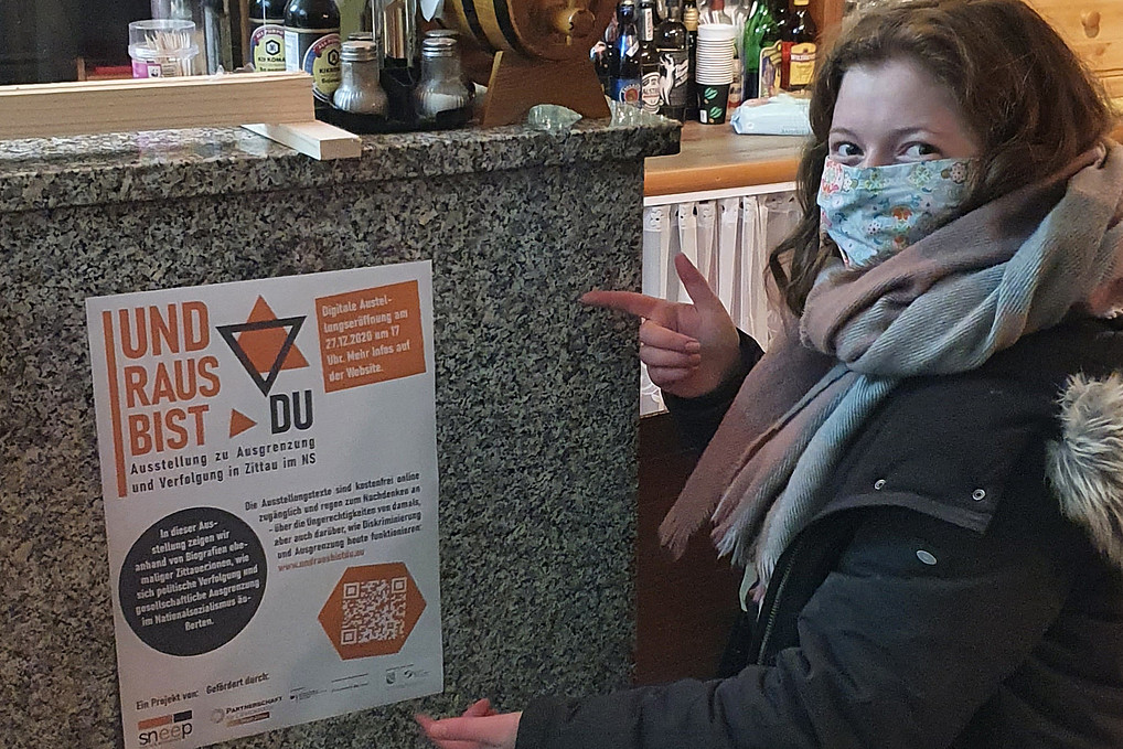 Nicola Bell steht in einer Studentenbar und zeigt auf das "Und raus bist du"-Plakat.