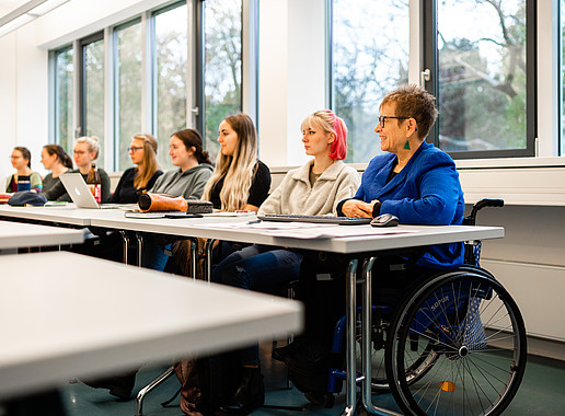 Seminarsituation mit mehreren Studierenden. Am Tischende sitzt die Professorin, die in einem Rollstuhl sitzt. 