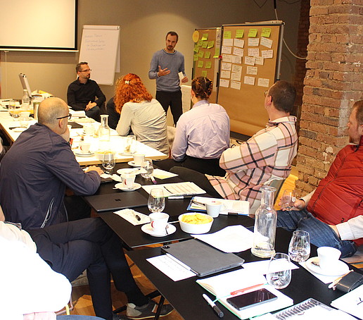 Workshop-Teilnehmende arbeiten an einem langen Tisch, im Hintergrund befindet sich eine Pinnwand mit angepinnten Karteikarten.