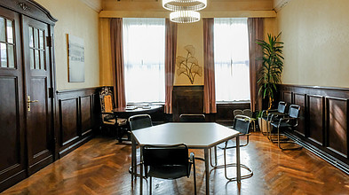 Ein historisch anmutender Raum, in dem ein sechseckiger Tisch mit Stühlen steht, an den Wänden sieht man Holzvertäfelung.