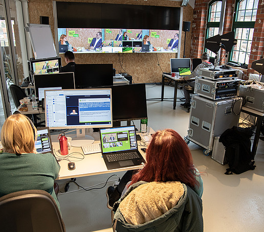 In einem großen Büroraum sind viele technische Geräte und Bildschirme zu sehen. Mitarbeitende sitzen an ihren Arbeitsplätzen und verfolgen den Live-Stream.