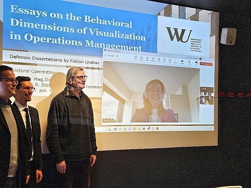 Three men are standing on the left in front of an illuminated projector screen on which Professor Keil can be seen.