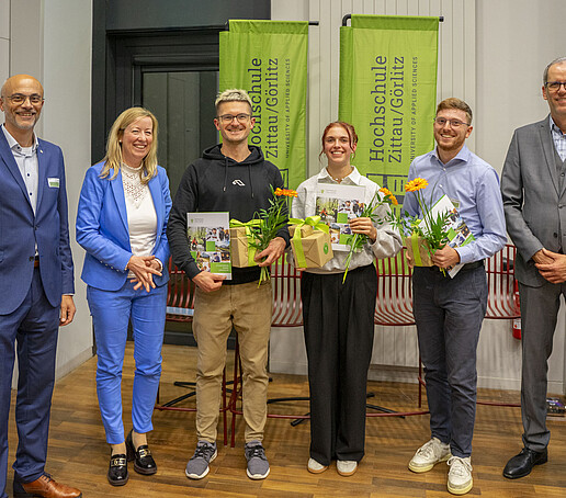 The award winners, Mr. Linack, Vice President Keil and Prof. Maiwald stand next to each other in front of green HSZG flags and smile into the camera.