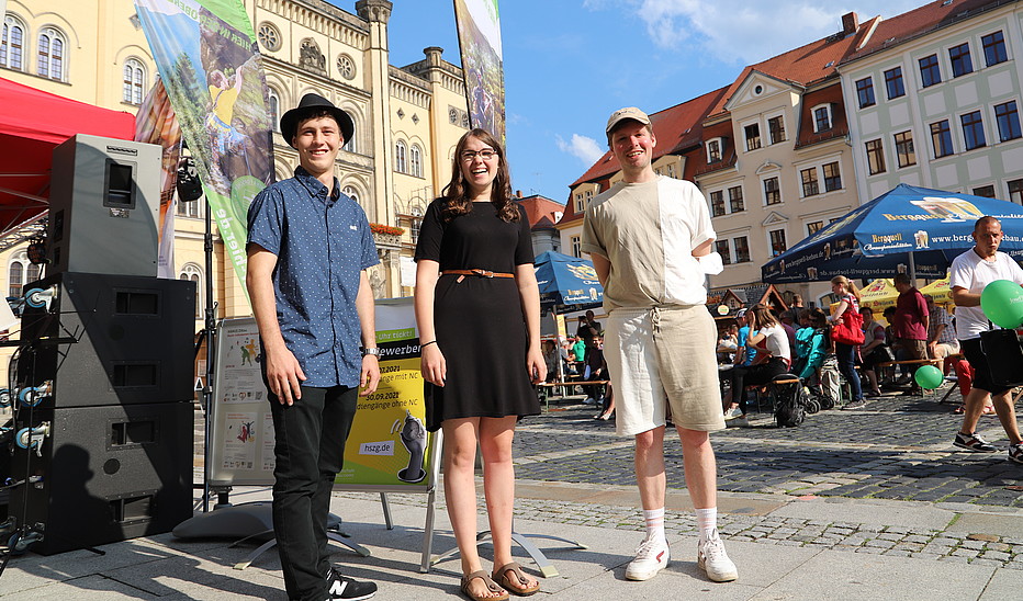 Foto: Susanne Fentzel Auf dem Zittauer Marktplatz neben einer Bühne stehen zwei Studenten und eine Studentin und lächeln in die Kamera.