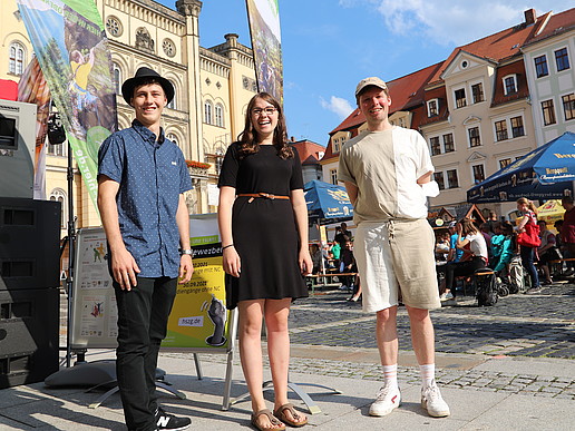 Two male and one female student are standing next to a stage on Zittau's market square, smiling into the camera.