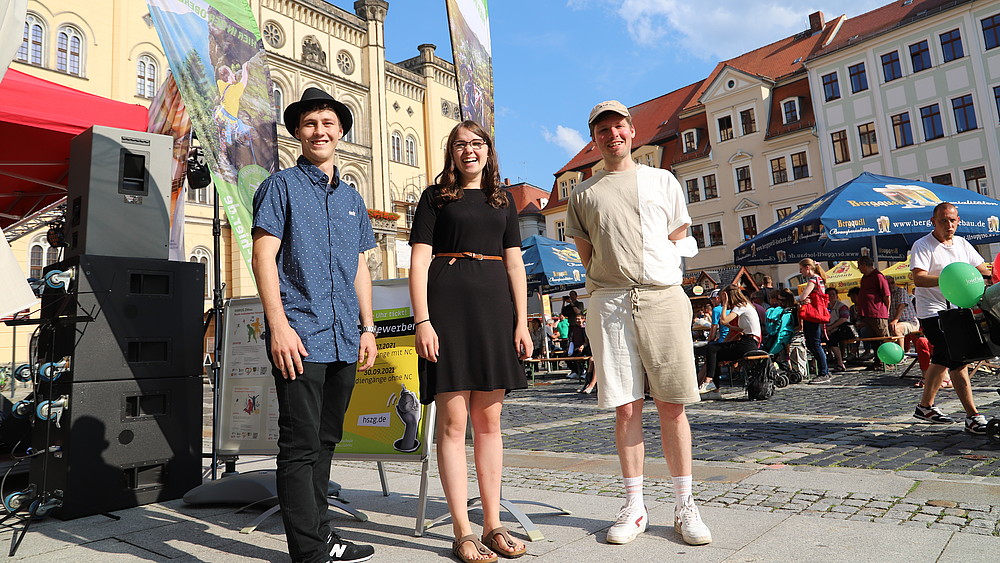 Two male and one female student are standing next to a stage on Zittau's market square, smiling into the camera.