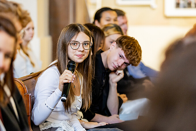 A young woman wearing glasses is sitting in a church pew and speaking into a microphone.