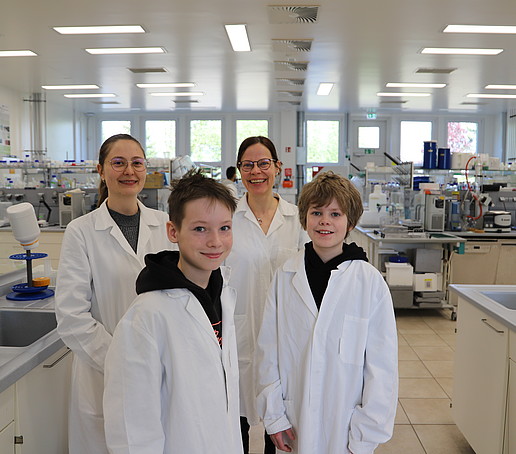 A lab technician, a professor and two students in white coats stand in the pharmaceutical biotechnology lab and smile at the camera.