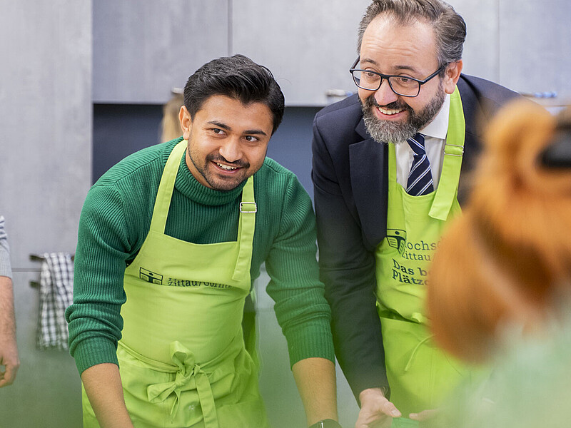 Minister of State and foreign student in aprons cut out cookies with green molds. They lean over the table and smile at the camera.