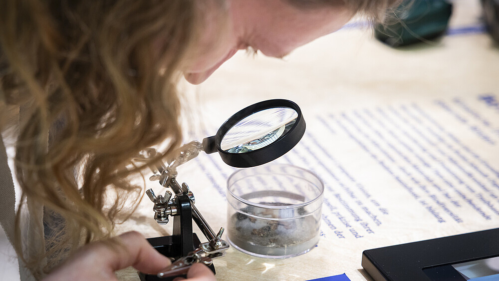A student looks through a magnifying glass into a glass dish filled with a sample.