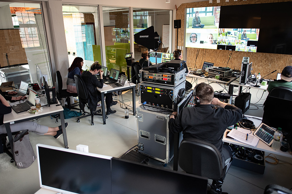 A broadcasting center with several desks and a large screen wall can be seen. Employees work at desks with their laptops.