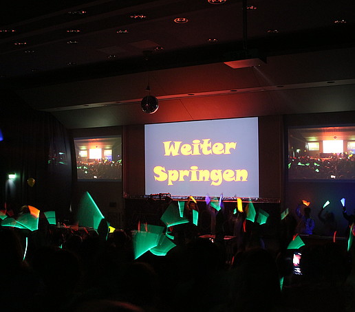 Green glow sticks light up in the dark Audimax and the projector screen says "Jump on".