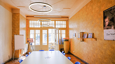 Zu sehen ist ein Seminarraum mit großem Tisch und Stühlen. Ein Poster von dem Film "Der Vorleser" hängt an der rechten Wand.