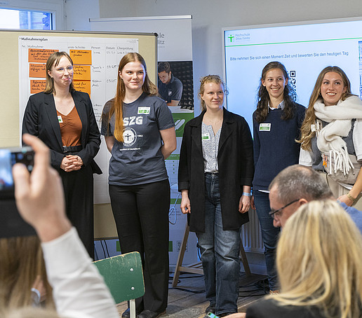 A group of four women stand in front of a full row of chairs. There is a pinboard in the background and a large screen with a presentation hanging on the wall.