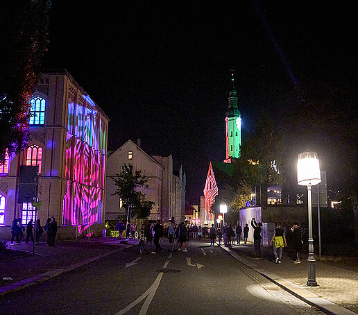 The monastery church is brightly lit in the background, to the left of it the staple gable is illuminated in purple.