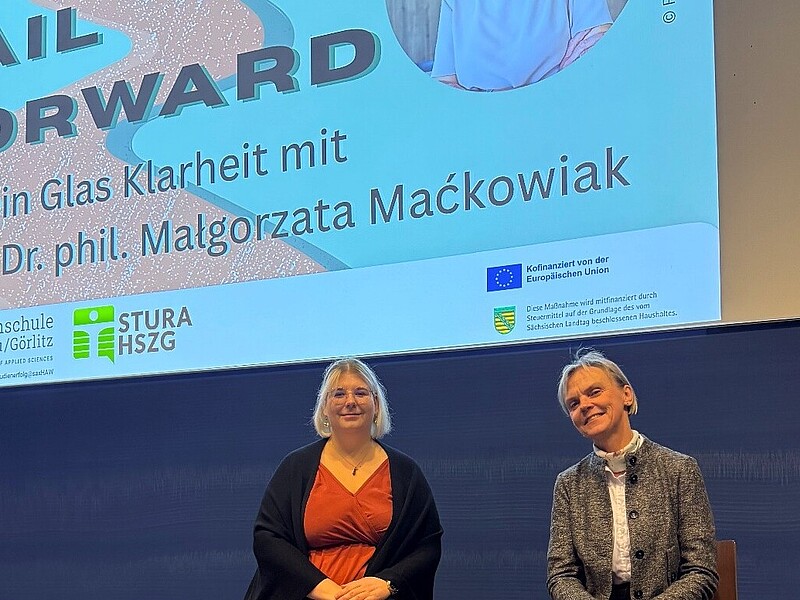 Aline Schulz and Vice President Mackowiak sit on high chairs in front of a blue lecture hall blackboard. A beamer projection with the headline "Fail Forward" appears above them.