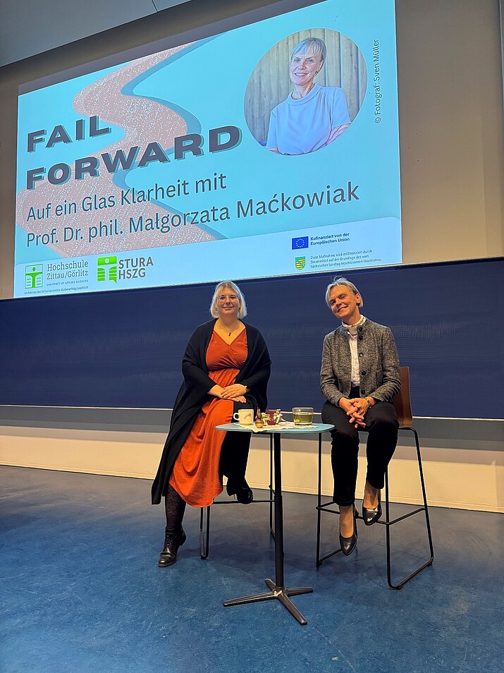 Two women sit on high chairs in front of a projector in the lecture hall.