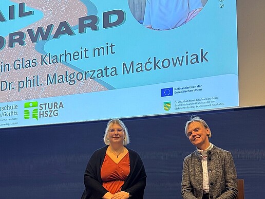 Aline Schulz and Vice President Mackowiak sit on high chairs in front of a blue lecture hall blackboard. A beamer projection with the headline "Fail Forward" appears above them.