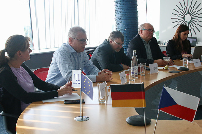Three men and two women are sitting at a table. The German and Czech flags are placed on the side of the table in the foreground as table decorations.