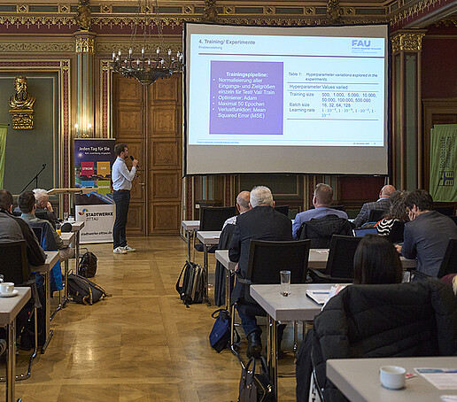 Ein Teilnehmer hält einen Vortrag und verweist auf die Leinwand mit digitaler Präsentation.