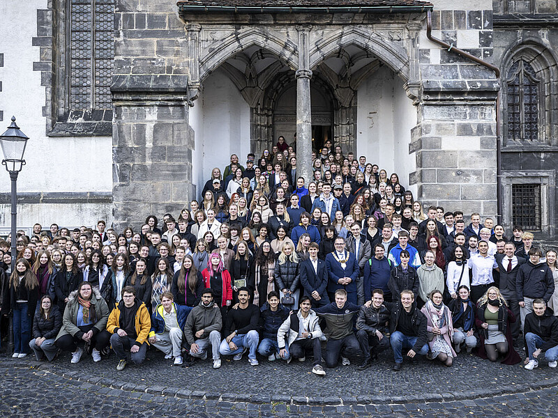 Eine große Menge junge Studierende und der Rektor stehen am Seiteneingang der Kirche auf den Stufen und dem vorderen Platz verteilt. Alle schauen in die Kamera.