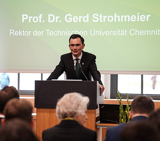 A man in a dark suit stands at a lectern and speaks to an audience. A screen with the inscription "Prof. Dr. Gerd Strohmeier, Rector of Chemnitz University of Technology" can be seen in the background.