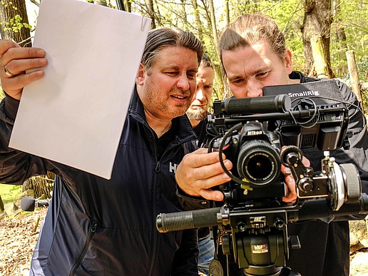 A cameraman stands behind a camera in the forest and adjusts it. An assistant is holding a white sheet for white balancing.